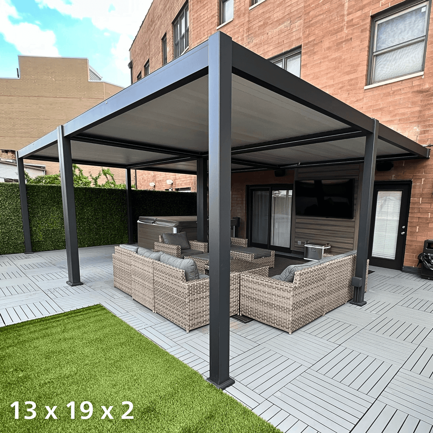 The black 13'x19' Bon Pergola features a retractable sunshade. The louvers on the top are fully closed, creating a sleek black roof. The structure is made of sturdy black columns and beams, with one side attached to the exterior wall of the house. The terrace beneath is furnished with outdoor furniture, including woven sofas and armchairs, with light gray cushions and pillows for added comfort.