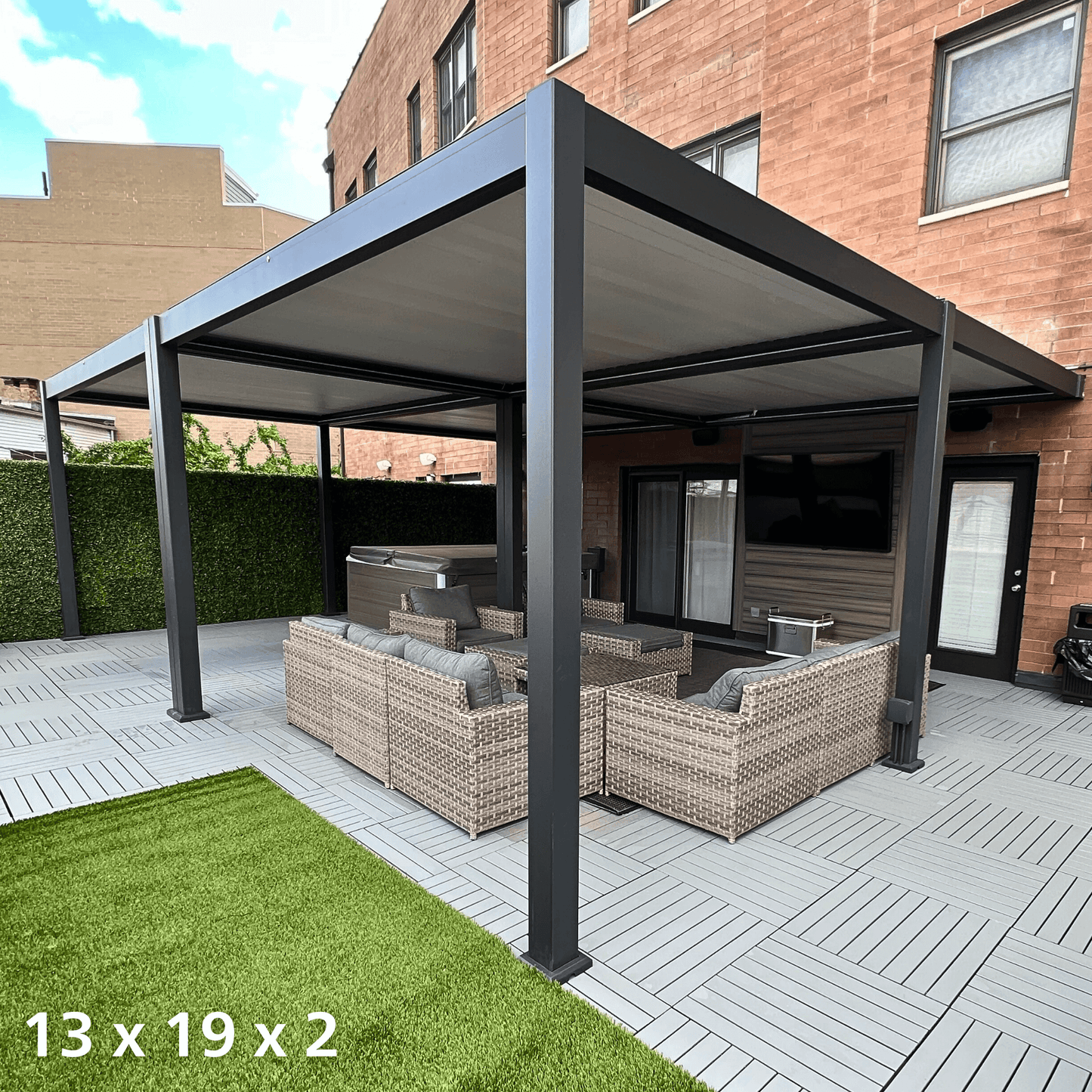 The black 13'x19' Bon Pergola features a retractable sunshade. The louvers on the top are fully closed, creating a sleek black roof. The structure is made of sturdy black columns and beams, with one side attached to the exterior wall of the house. The terrace beneath is furnished with outdoor furniture, including woven sofas and armchairs, with light gray cushions and pillows for added comfort.