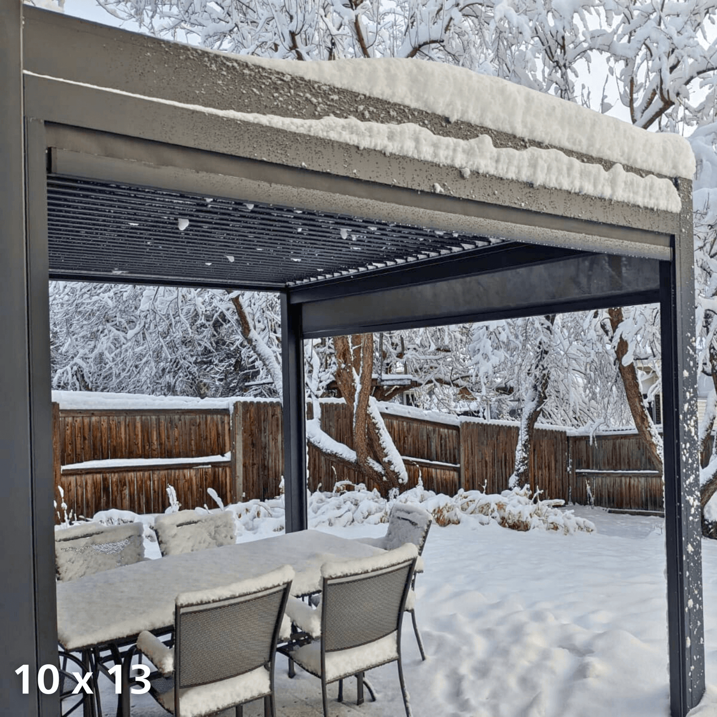 The dark Bon Pergola 10x13 electric louvered pergola features louvers partially open, covered in a thick blanket of white snow. Its structure, made of dark gray columns and beams, is also coated with snow. Below, an outdoor dining set consisting of a rectangular table and chairs is completely covered in snow, creating a serene winter landscape of silver and white.
