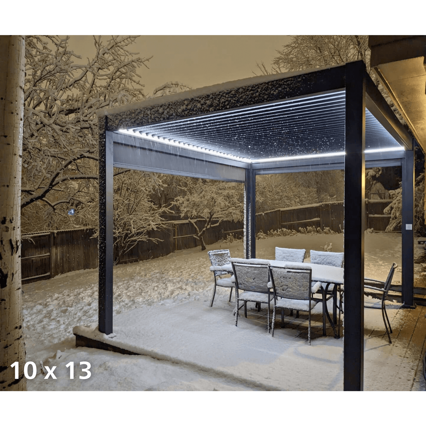 An outdoor terrace covered in thick snow features a 10x13 deep gray Bon Pergola with motorized louvers. The louvers are partially open, with snow accumulating on top. The structure consists of deep gray posts and beams, and the edges of the beams are adorned with white LED light strips. These lights illuminate the area below the pergola and highlight the gently falling snowflakes, creating a serene and magical winter atmosphere.