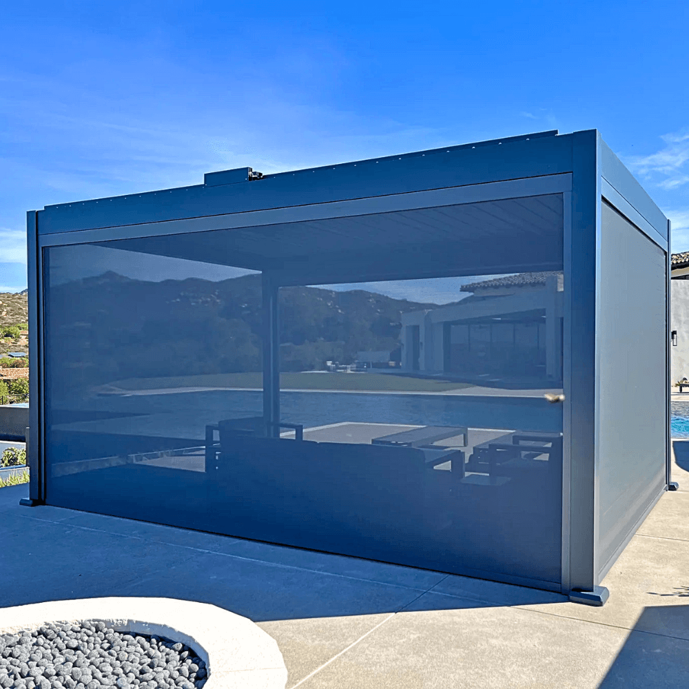 The dark gray Bon Pergola is equipped with wind-resistant sunshades on all four sides. These shades are mostly lowered, creating a more private space while blocking wind and sunlight. The shades are designed to offer wind protection, UV defense, and some waterproofing, making the pergola a versatile outdoor space that can be enjoyed in any weather.