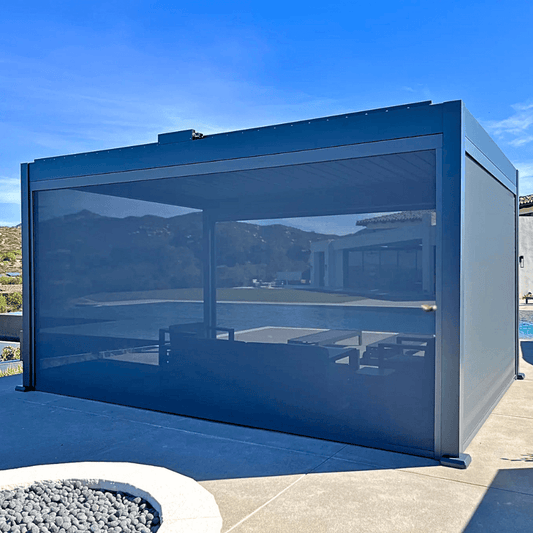 The dark gray Bon Pergola is equipped with wind-resistant sunshades on all four sides. These shades are mostly lowered, creating a more private space while blocking wind and sunlight. The shades are designed to offer wind protection, UV defense, and some waterproofing, making the pergola a versatile outdoor space that can be enjoyed in any weather.