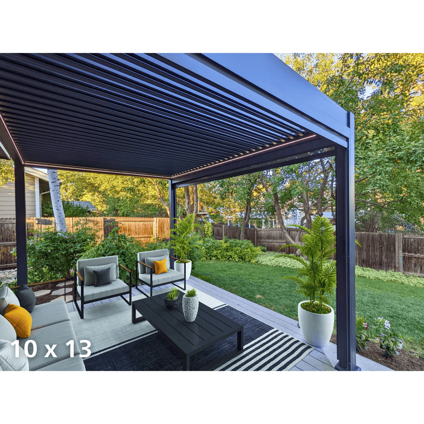The black Bon Pergola features partially open louvers, casting slender, striped shadows below. Its structure consists of sturdy black columns and beams, with the beam edges illuminated by warm yellow LED strip lights, creating a cozy atmosphere. Beneath it sits a modern outdoor seating area with a light gray L-shaped sofa, two armchairs with light gray cushions, and a black rectangular coffee table. The pergola is surrounded by green grass, shrubs, and trees, adding to the tranquil outdoor setting.