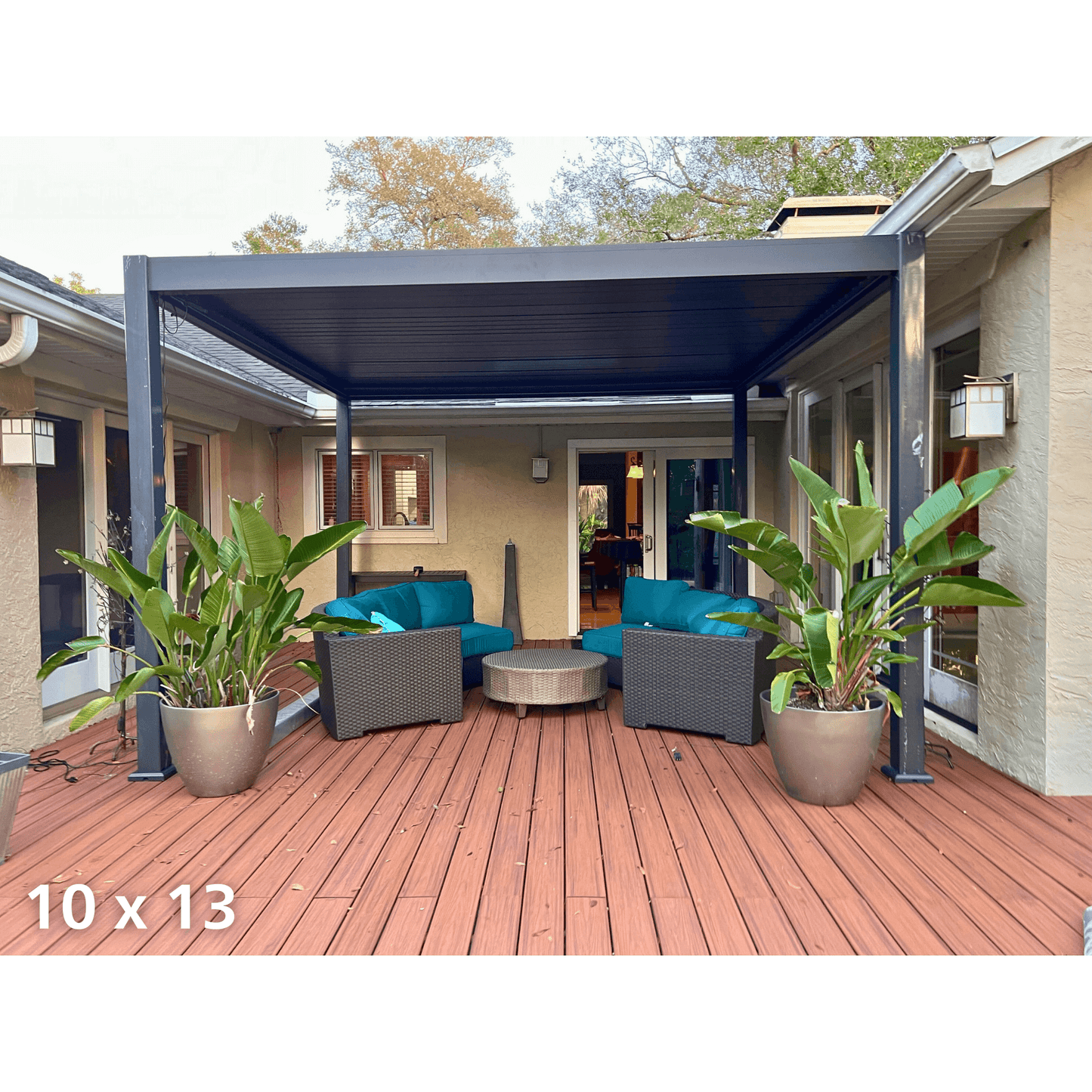 The dark gray Bon Pergola features louvers in a closed position, creating a solid gray roof that provides full shade. The structure is supported by thick gray columns and beams. Below, a patio with reddish-brown wooden flooring is furnished with outdoor lounge furniture, including two dark gray woven armchairs with vibrant teal cushions and pillows, as well as a round light gray woven coffee table.