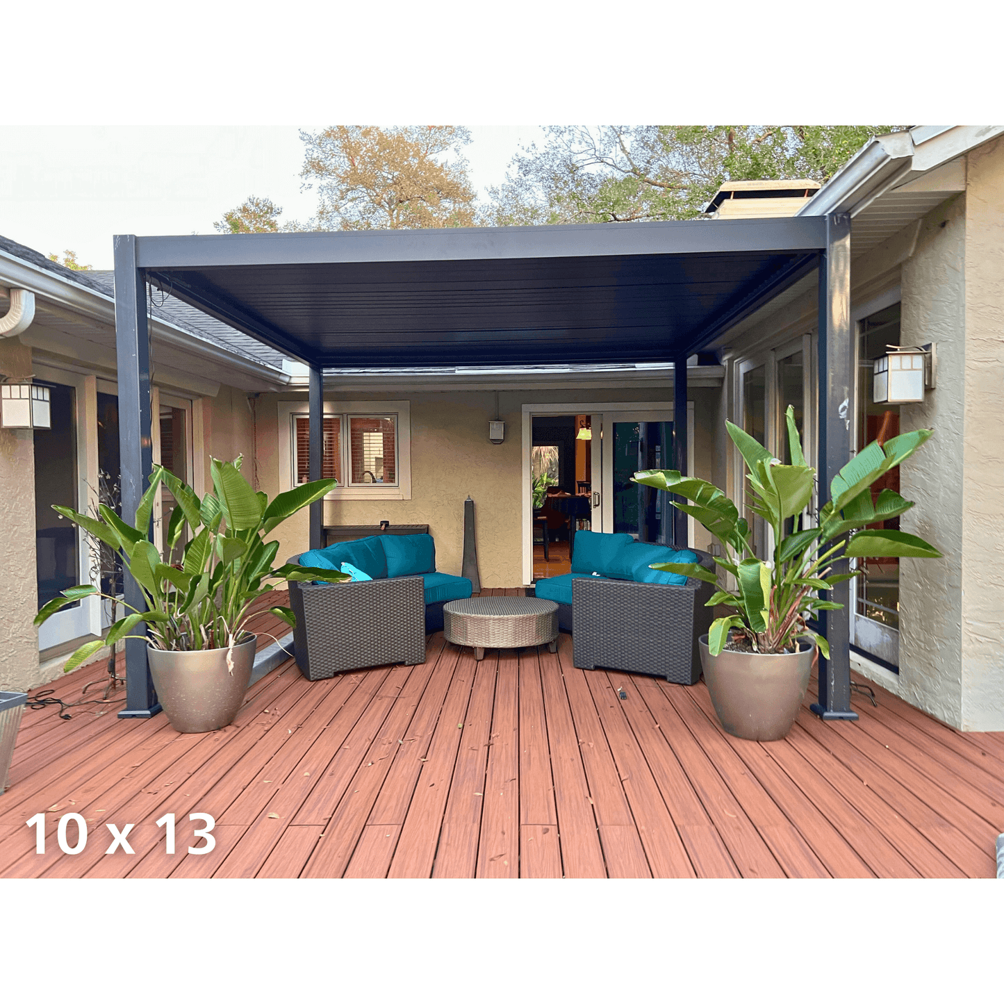 The dark gray Bon Pergola features louvers in a closed position, creating a solid gray roof that provides full shade. The structure is supported by thick gray columns and beams. Below, a patio with reddish-brown wooden flooring is furnished with outdoor lounge furniture, including two dark gray woven armchairs with vibrant teal cushions and pillows, as well as a round light gray woven coffee table.