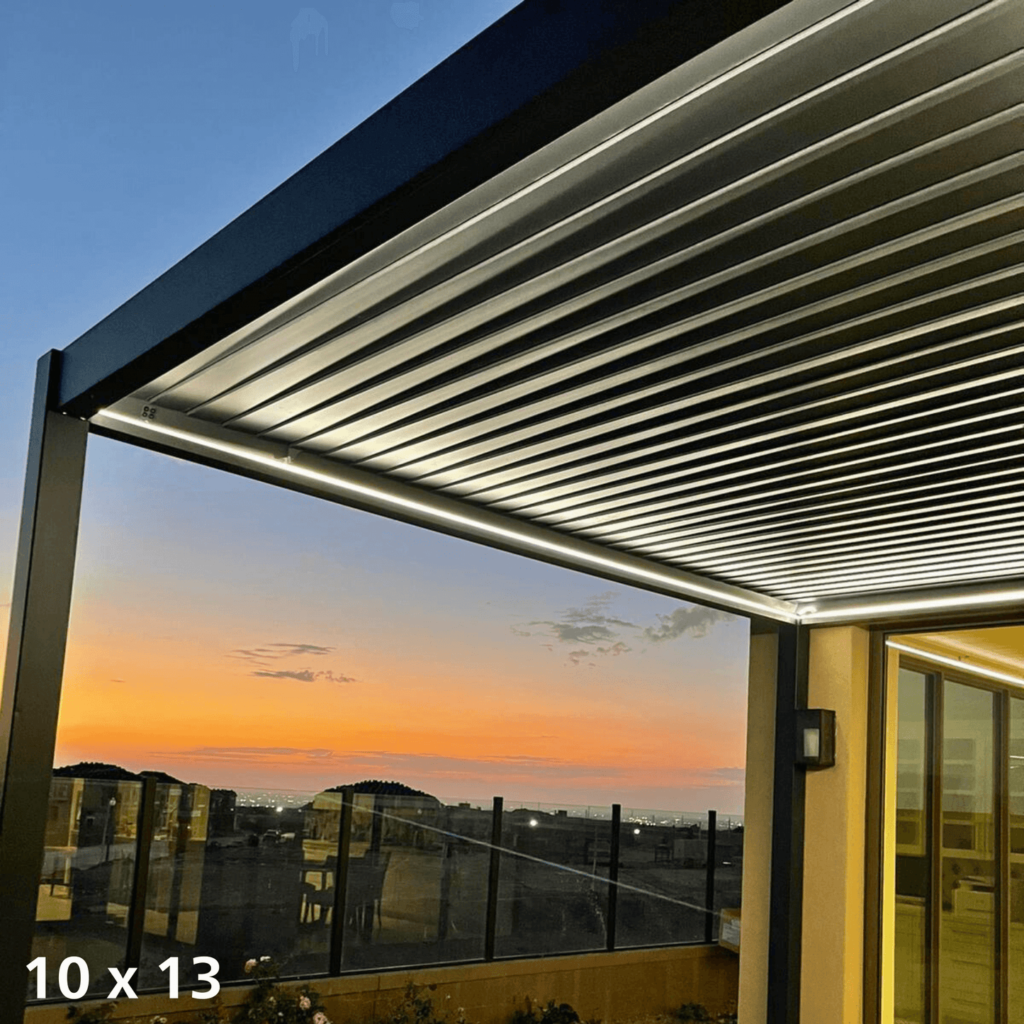The dark-colored electric pergola features a sleek structure with dark posts and beams. Along the edges of the top and beams, a warm yellow LED light strip provides soft illumination, casting a gentle glow beneath. On one side, an opening reveals a stunning sunset view, with distant buildings clearly outlined against the vibrant sky. This creates a cozy and inviting atmosphere, perfect for relaxing while enjoying the evening scenery.