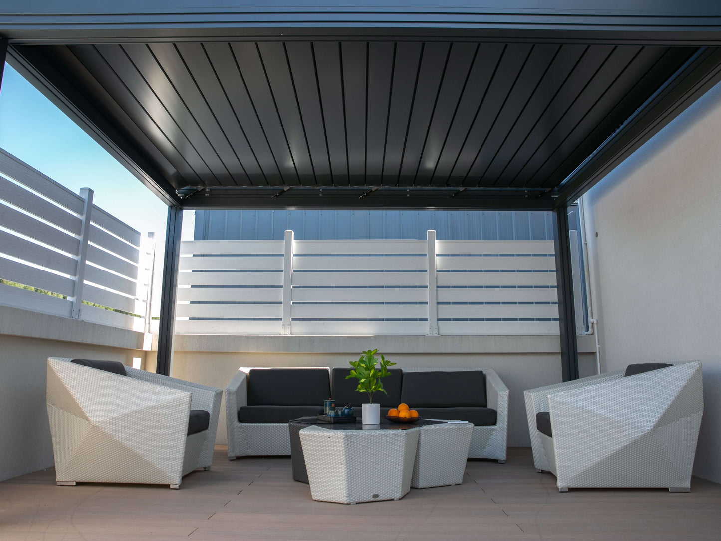 Beneath the dark gray pergola is a patio paved with light-colored tiles, furnished with a white wicker outdoor set. It includes a sofa and two armchairs with dark gray cushions, along with a white geometric coffee table. The white patio, furniture, and railing contrast beautifully with the dark pergola, creating a bright, comfortable outdoor lounge space.