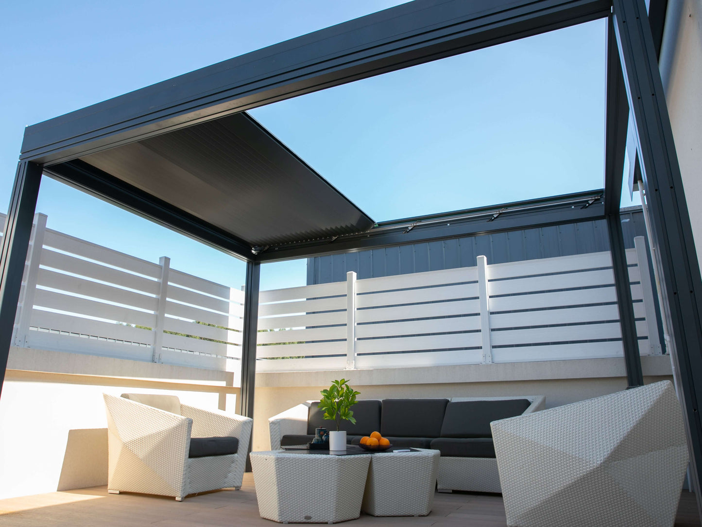 The pergola’s roof is partially open, letting sunlight stream through. Below is a white patio furnished with a white wicker outdoor set, including a sofa and two armchairs with dark gray cushions, and a white coffee table. On the table sits a potted plant and a few pieces of fruit. The patio is enclosed by a white horizontal slat railing, and the open louvers above reveal a view of the bright blue sky.