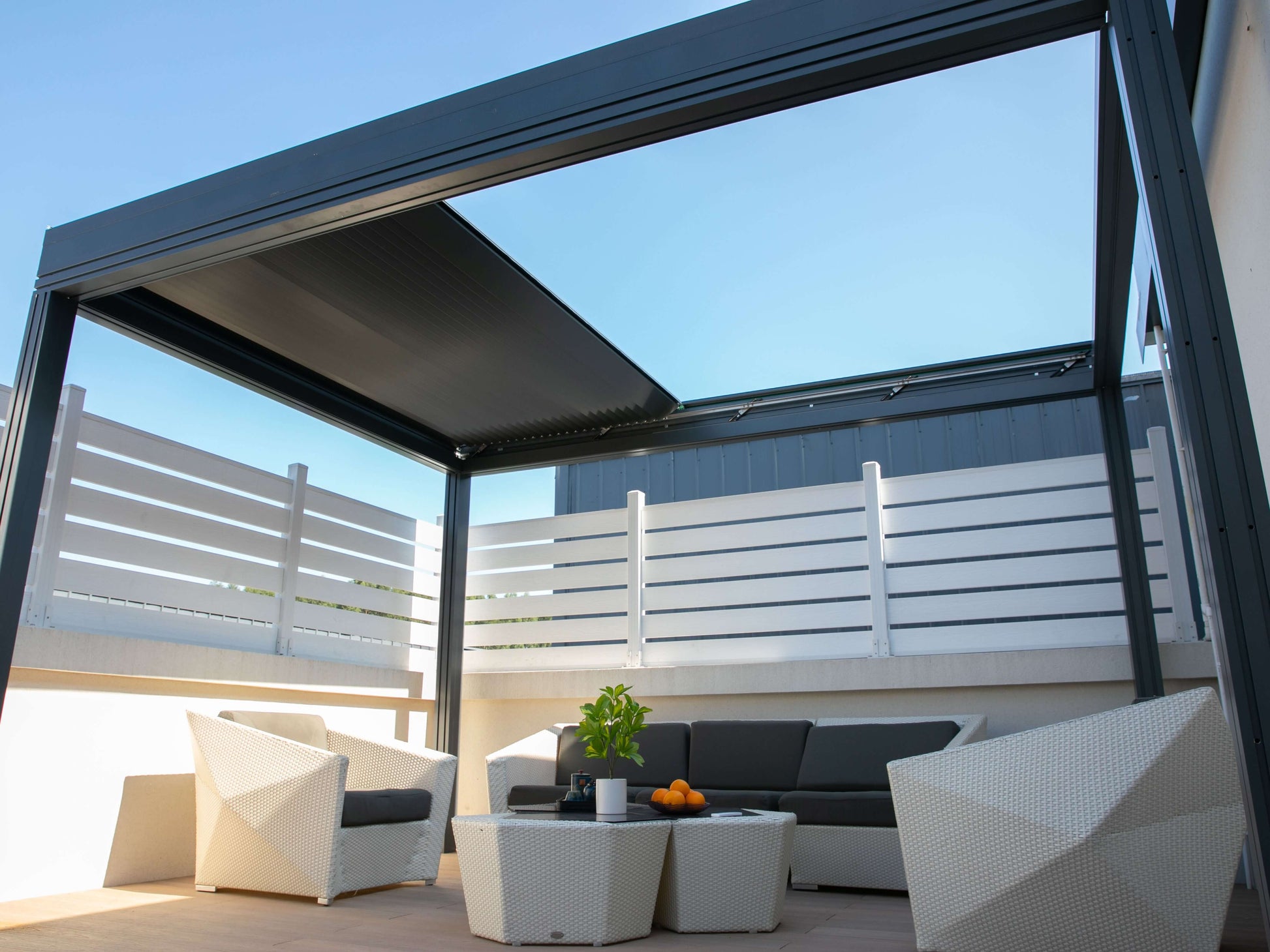 The pergola’s roof is partially open, letting sunlight stream through. Below is a white patio furnished with a white wicker outdoor set, including a sofa and two armchairs with dark gray cushions, and a white coffee table. On the table sits a potted plant and a few pieces of fruit. The patio is enclosed by a white horizontal slat railing, and the open louvers above reveal a view of the bright blue sky.