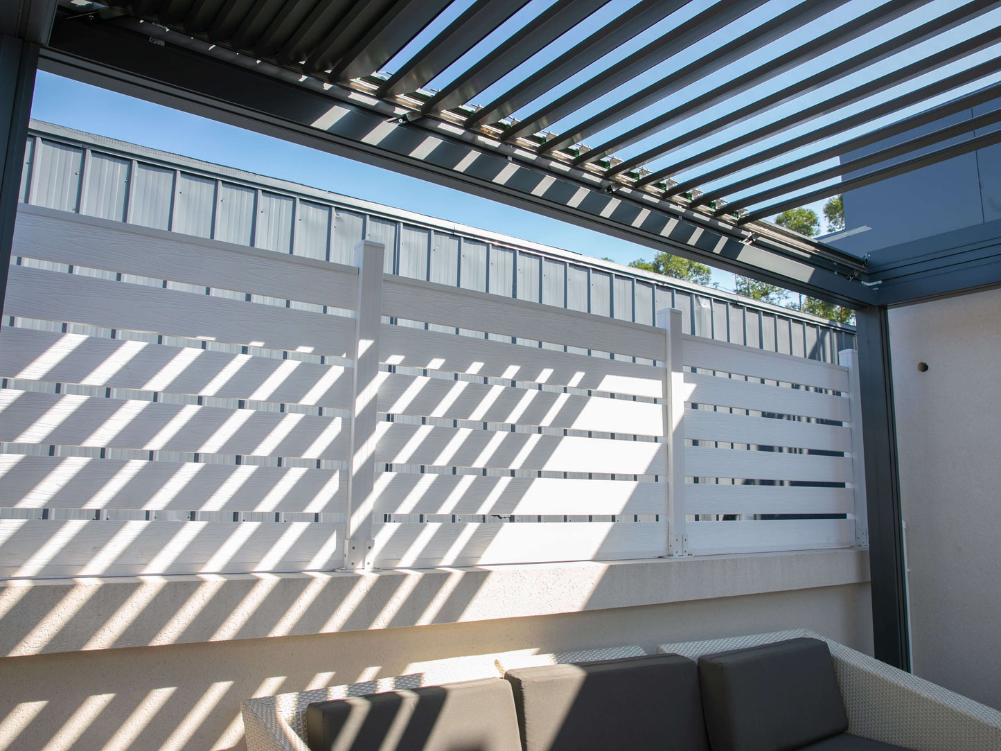 The dark gray pergola features partially open louvers, with sunlight filtering through the deep gray slats. Its structure is made up of matching dark gray beams. Along the sides, white horizontal slat railings add contrast, casting angled shadows as sunlight passes through the gaps, creating a dynamic and inviting outdoor atmosphere.