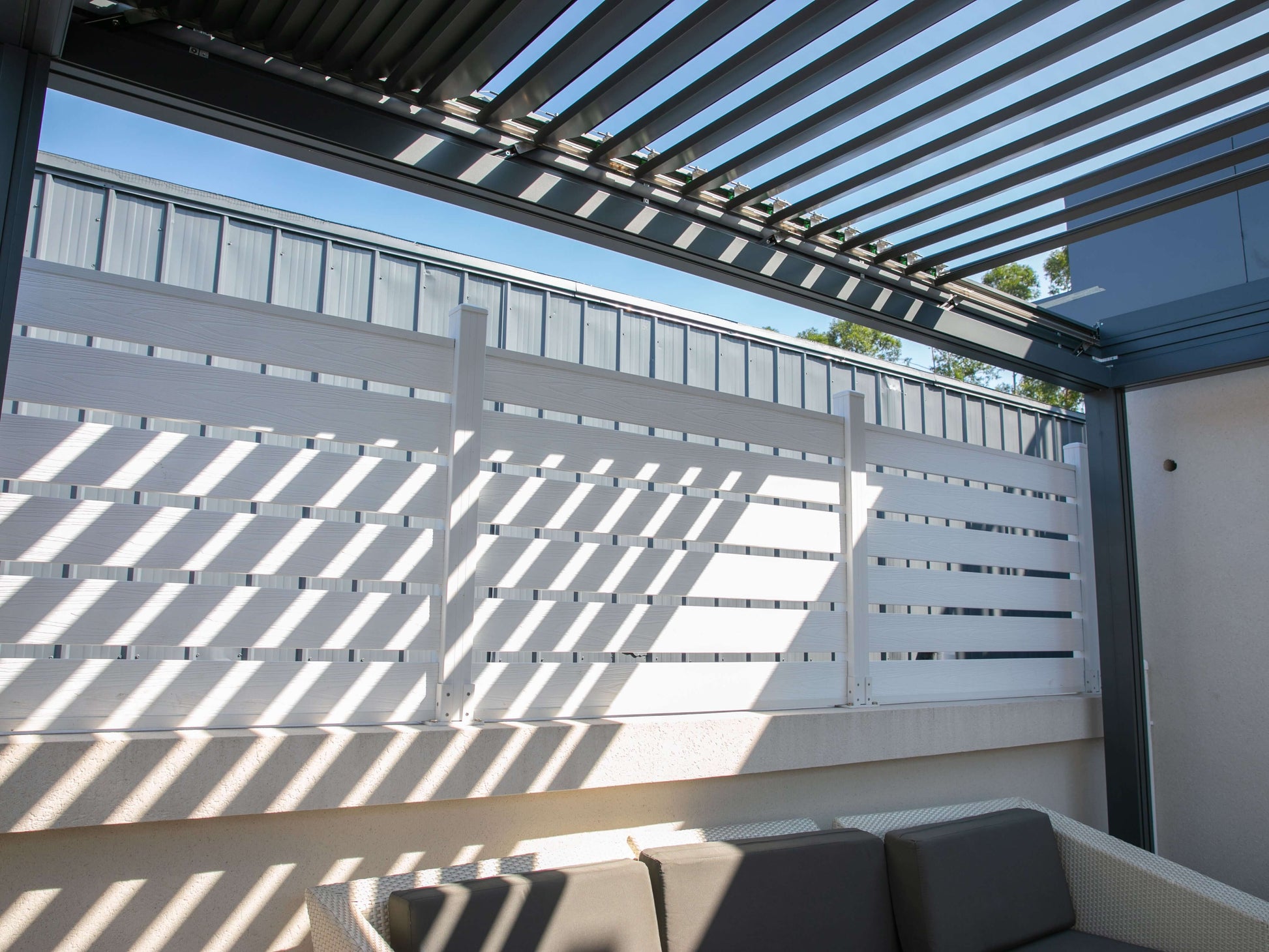 The dark gray pergola features partially open louvers, with sunlight filtering through the deep gray slats. Its structure is made up of matching dark gray beams. Along the sides, white horizontal slat railings add contrast, casting angled shadows as sunlight passes through the gaps, creating a dynamic and inviting outdoor atmosphere.