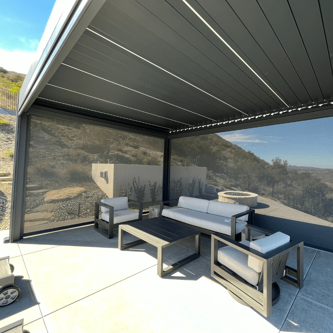 This is a dark gray Bon Pergola with a clearly visible motorized louvered roof. The structure is built with sturdy dark gray posts and beams. On the sides, dark shade screens provide coverage for the modern outdoor lounge furniture beneath. Through the partially lowered screens, you can catch glimpses of distant hills and surrounding greenery.