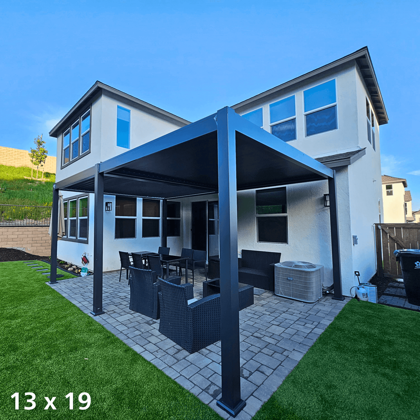 This daytime photo shows a black 13'x19' Bon Pergola installed on the backyard terrace, providing full shade. The pergola's structure is made of sturdy black columns and beams, with one side attached to the exterior wall of the house. Beneath it, outdoor furniture is arranged, including a black rectangular dining table with matching chairs, along with a deep gray outdoor sofa and armchairs.