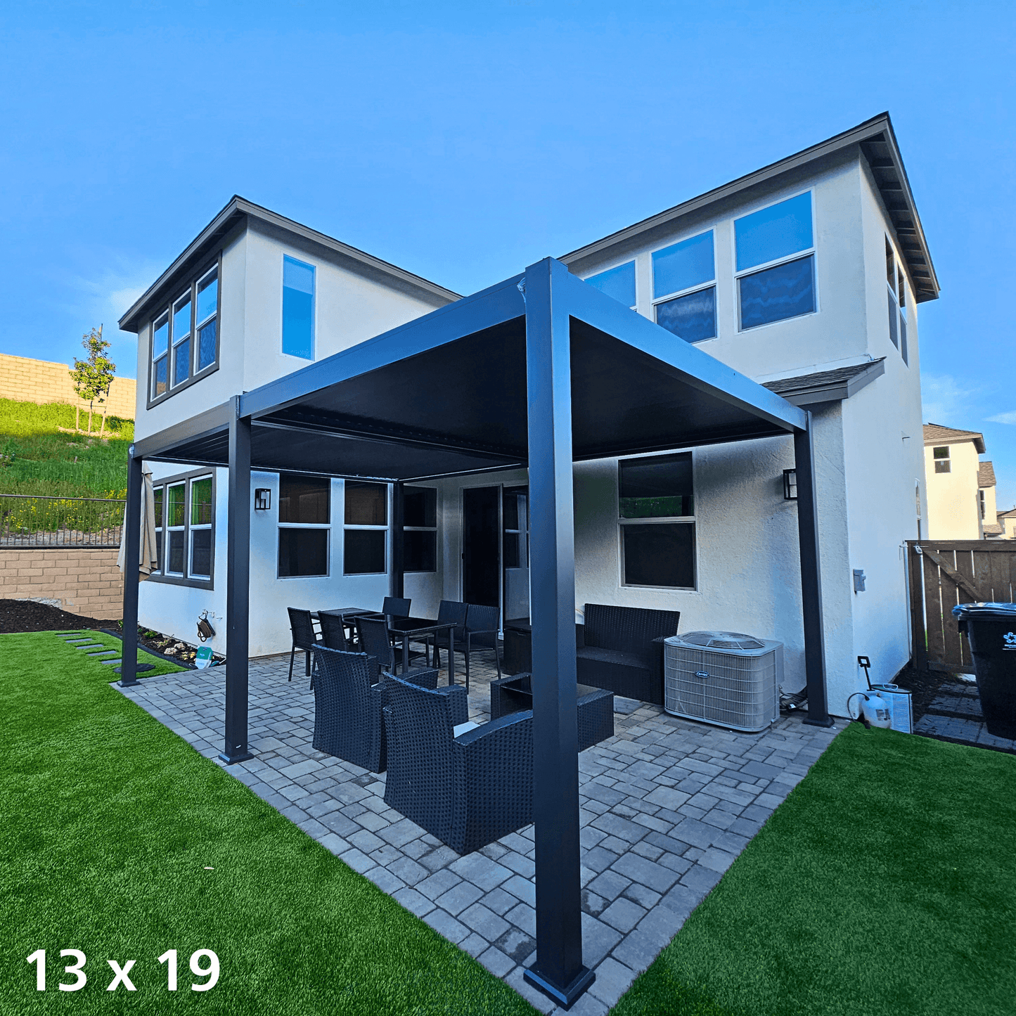 This daytime photo shows a black 13'x19' Bon Pergola installed on the backyard terrace, providing full shade. The pergola's structure is made of sturdy black columns and beams, with one side attached to the exterior wall of the house. Beneath it, outdoor furniture is arranged, including a black rectangular dining table with matching chairs, along with a deep gray outdoor sofa and armchairs.