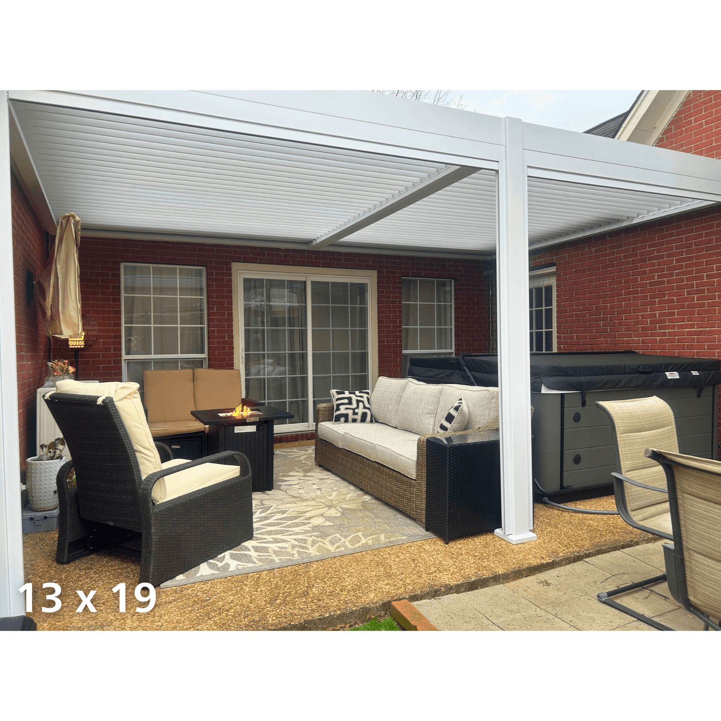 A white 13'x19' wall-mounted Bon Pergola is installed, with the louvers closed, creating a solid white roof. One side of the pergola is fixed to the red brick exterior wall of the house, while the other side is supported by white columns. The terrace is furnished with outdoor lounge furniture, including woven armchairs and a sofa with light-colored cushions and pillows. In the center of the seating area is a black square gas fire pit coffee table, with flames burning inside.