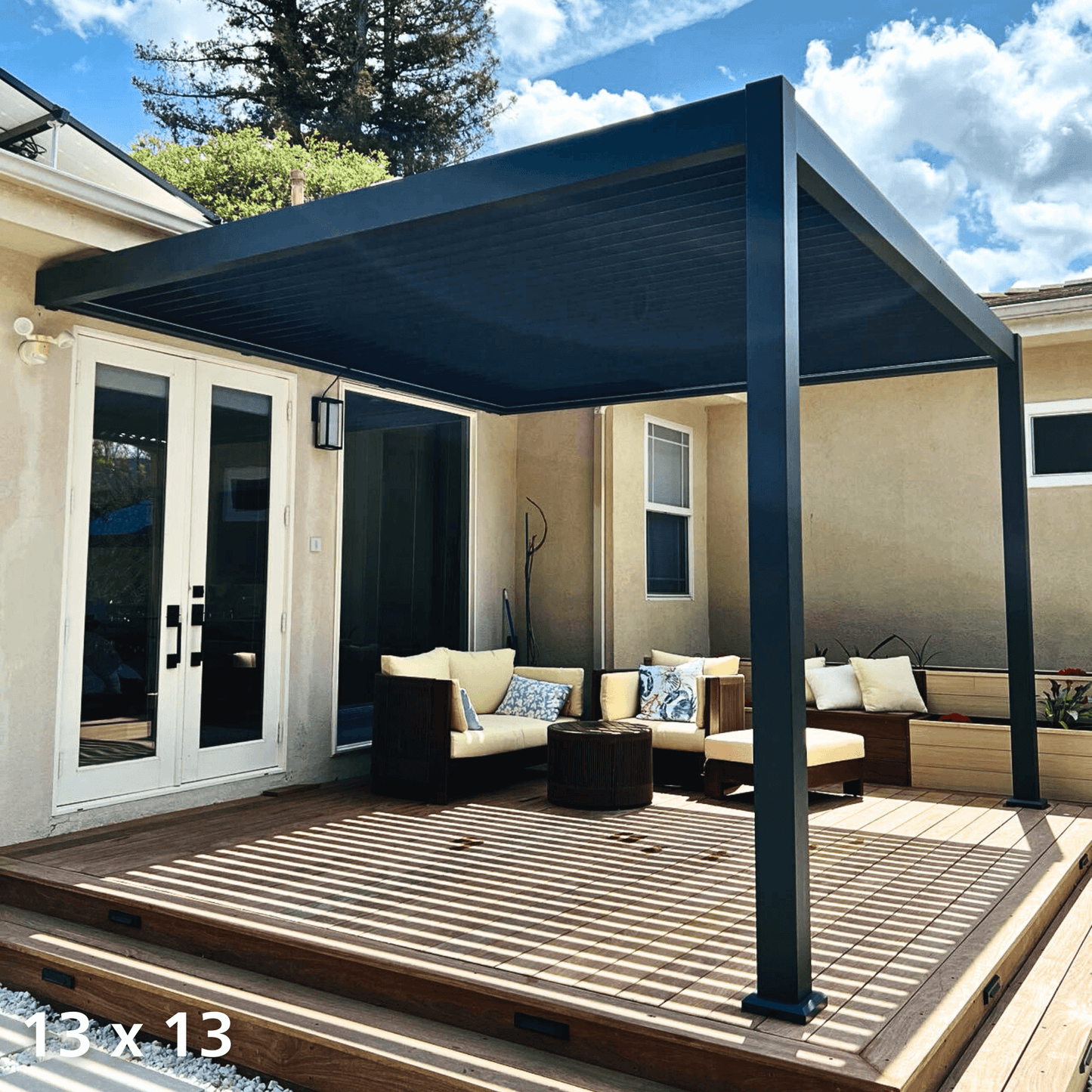 The black Bon Pergola is shown with its louvers fully closed, creating a solid deep gray roof that offers full shade. Its structure features bold, modern dark gray posts and beams, with one side attached to the home's exterior wall. Below is a wood-decked patio furnished with light-toned outdoor seating, including a sofa and armchairs, blending comfort with sleek, contemporary style.