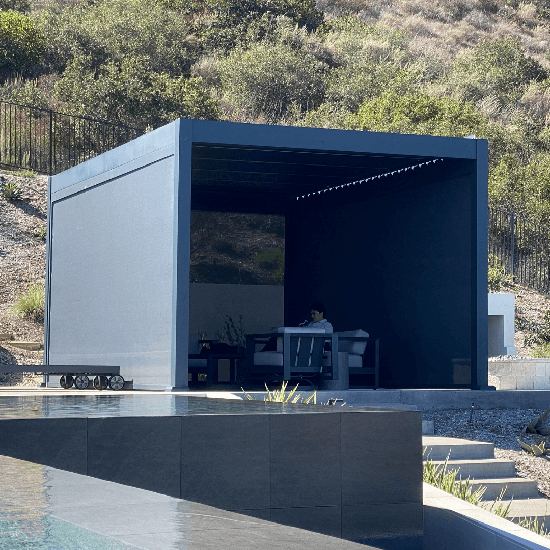 The dark gray Bon Pergola features closed louvers, creating a solid roof that provides full shade. Its sturdy frame is made of thick dark gray beams and posts, with windproof shade screens installed on both sides. Someone is seated inside, enjoying the sheltered space. The pergola sits on a platform with light gray flooring, positioned near the edge of a sleek black water feature or a low-profile pool.