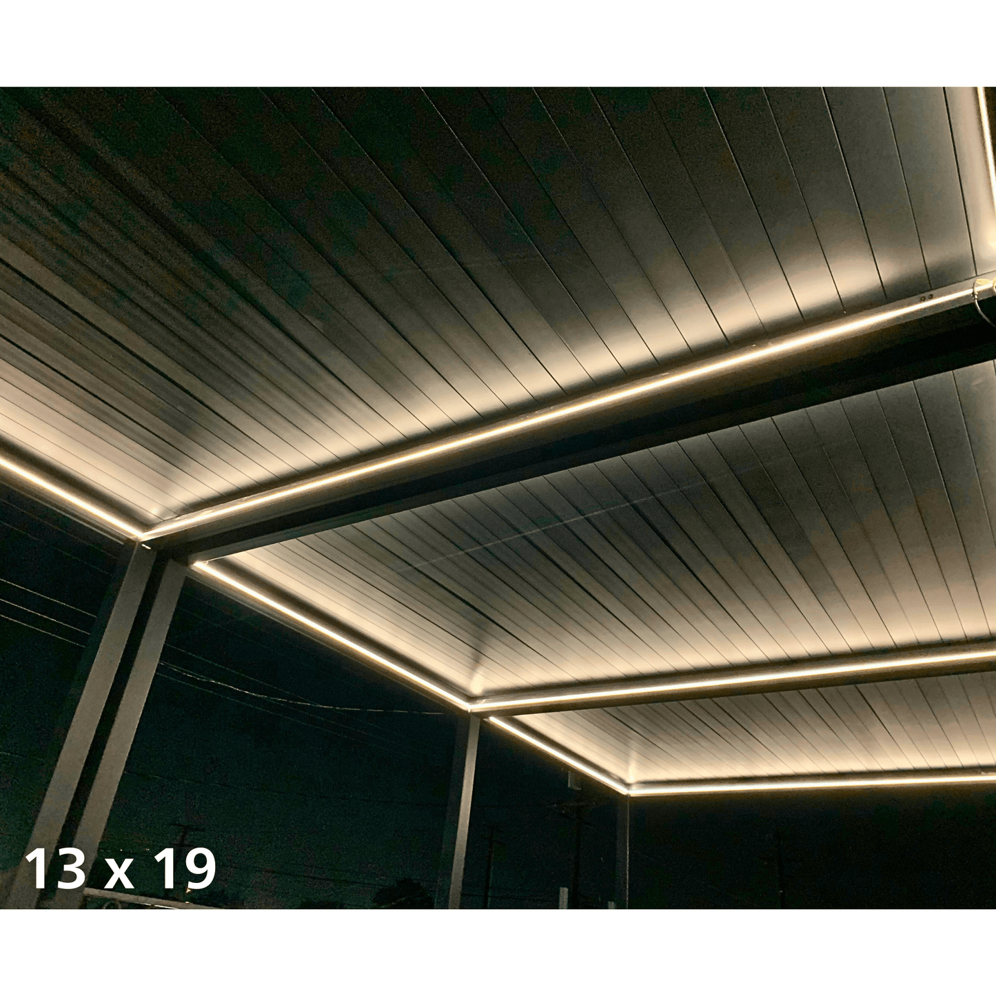 The top of the pergola features dark louvers in a closed position, forming a solid roof. The structure is made of dark columns and beams. Along the edges of the pergola’s top and beams, a warm yellow LED strip is installed, casting a soft glow upward onto the underside of the louvers. This creates a cozy, inviting atmosphere while also highlighting the pergola's outline.