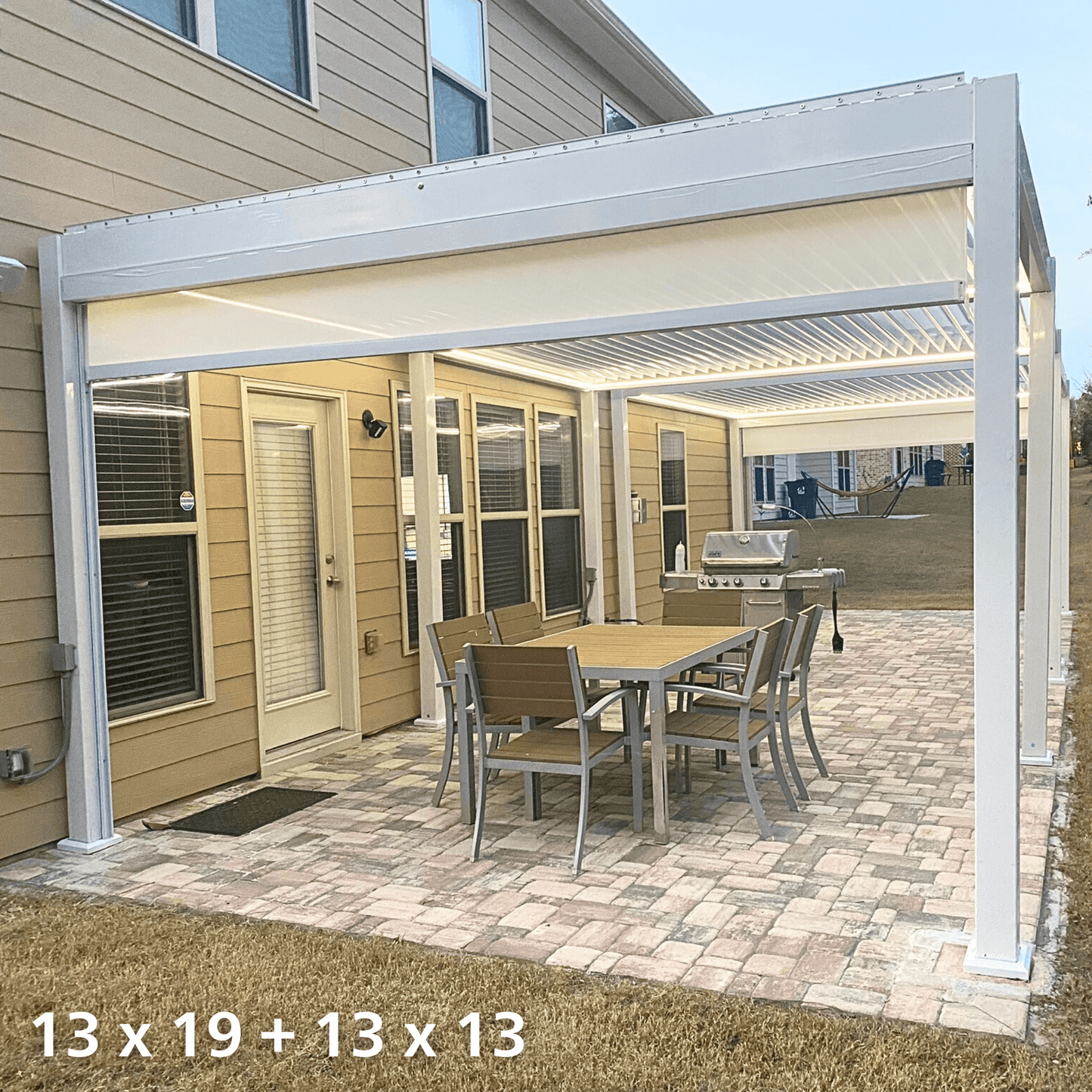 The white 13'x19' Bon Pergola with electric louvers is extended and connected to a smaller 13'x13' pergola of the same style. The louvers on both pergolas are partially open, showcasing their slatted structure. The pergola’s frame is made of white columns and beams, with one side attached to the exterior wall of the house. Outdoor furniture is placed beneath the pergolas, and white electric shading blinds are installed along the edges for additional sun protection.