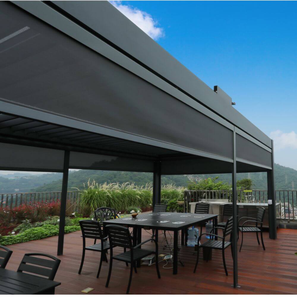 The dark gray Bon Pergola features dark windproof shade screens on at least two sides, creating a spacious outdoor dining or lounging area. The screens are partially lowered, providing shade, privacy, and wind protection. The terrace is furnished with dark outdoor dining chairs and a table, forming an outdoor dining zone. The edge of the terrace is equipped with metal railings, offering views of the distant greenery, hills, and a partial cityscape beyond.