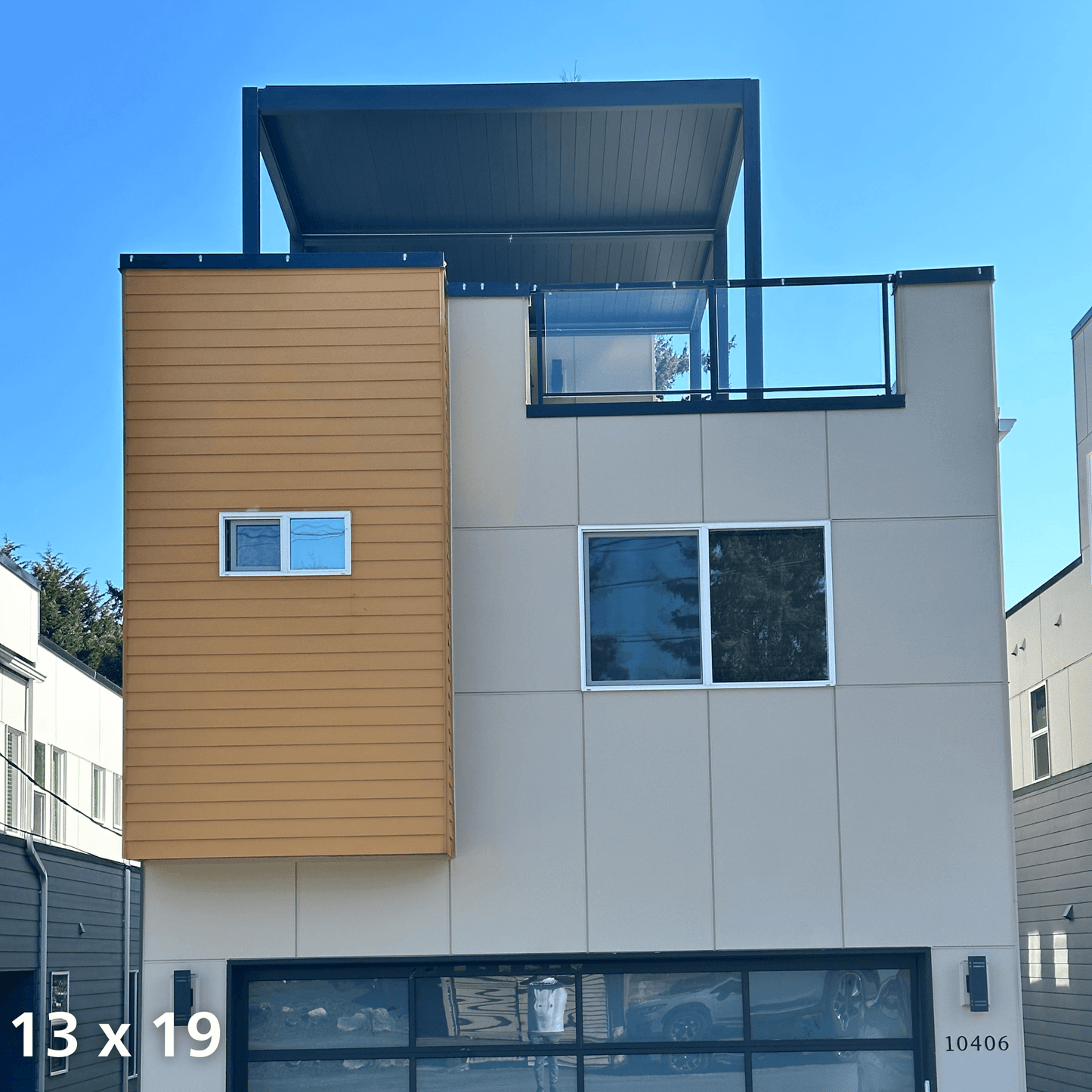 A 13'x19' dark gray Bon Pergola with motorized louvers sits on a rooftop terrace, its louvers partially open. The pergola features sleek dark gray posts and beams, complementing the modern design. Glass railings line the terrace, offering unobstructed views and a stylish, open feel.