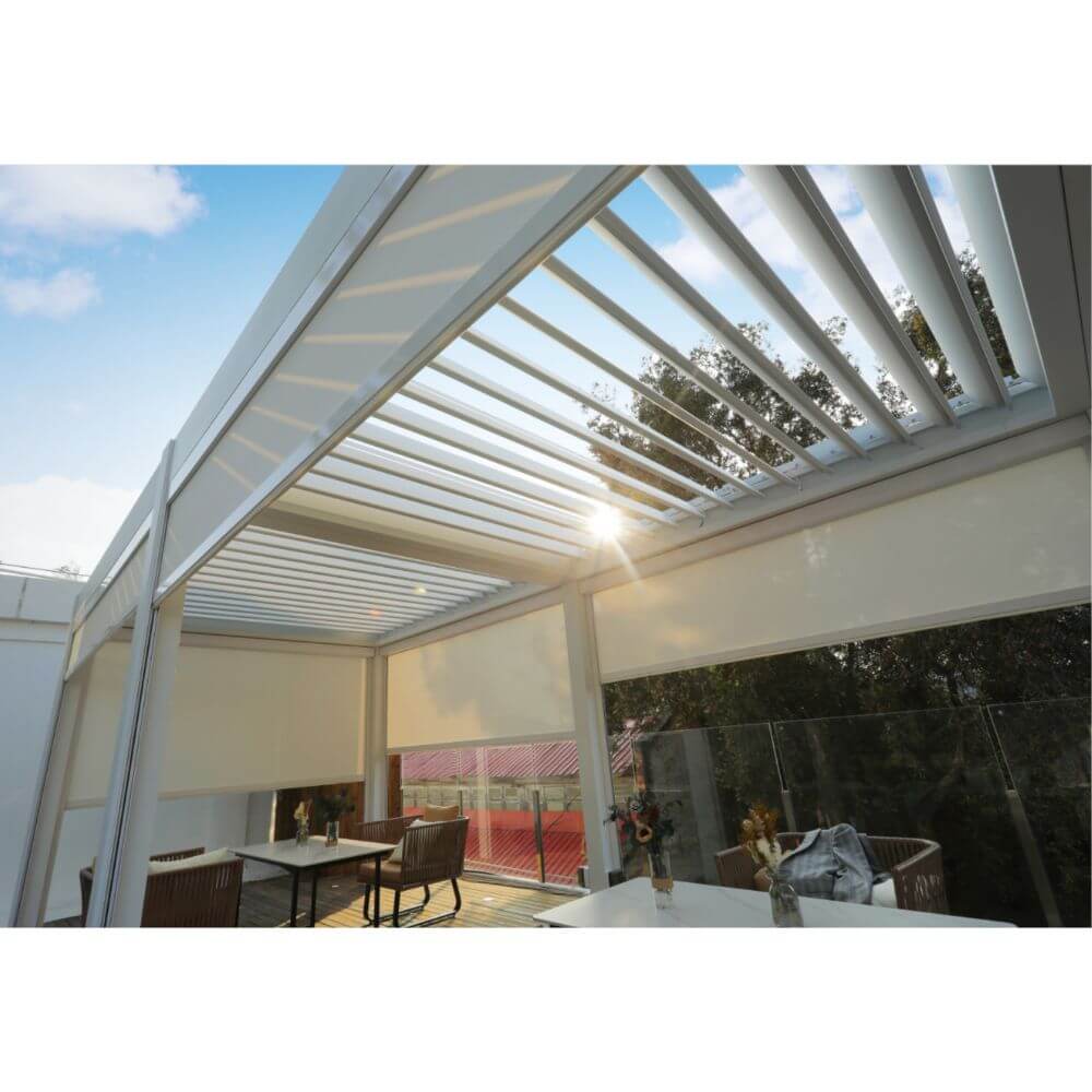 The white Bon Pergola features a partially open white louvered roof, with sunlight streaming through the gaps between the louvers. The structure is clearly visible, including white beams and the partially opened louvers. This adjustable louver system allows users to control the angle of the louvers, adjusting the amount of shade and light that enters the space, providing optimal sun control.