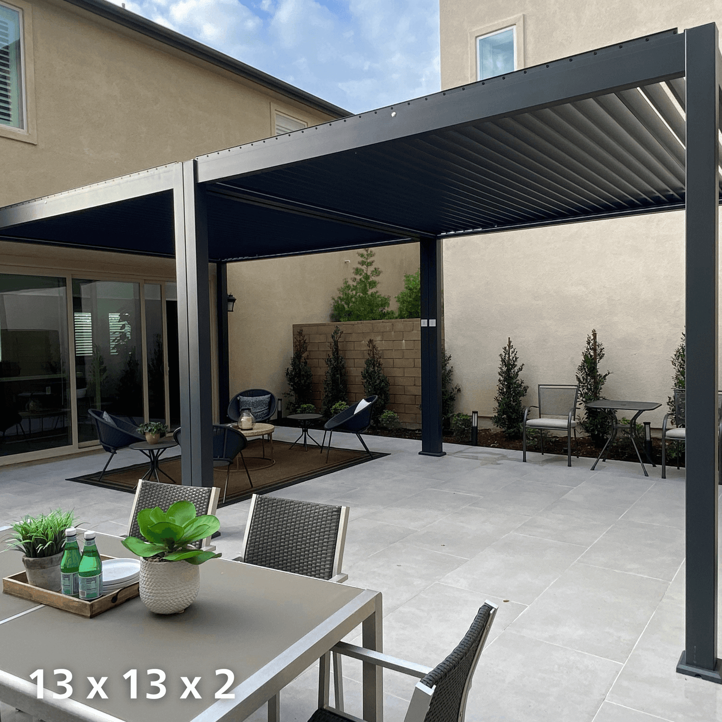 A black Bon Pergola 13'x13' with motorized louvers is installed above an outdoor terrace. The louvers are partially open, casting elongated, striped shadows on the ground. The pergola’s structure consists of sturdy black posts and beams, giving it a modern, minimalist look. Beneath the pergola, a light gray tiled terrace is divided into different relaxation zones, creating a versatile and stylish outdoor space.
