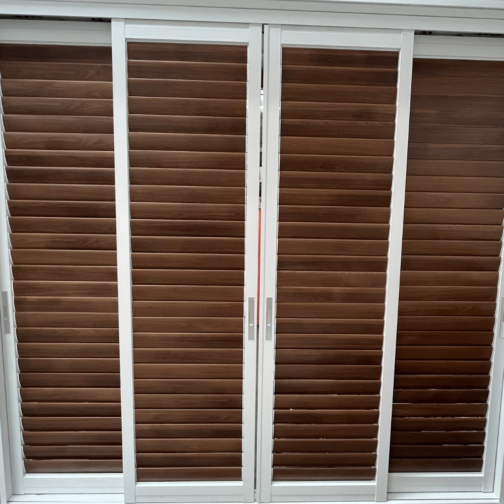 The sliding blinds feature white frames with deep brown horizontal slats. The slats have a wood-like texture, with a smooth, glossy finish and a uniform color. The two central panels are adjacent to each other and come with vertical silver handles, indicating they can slide open. The doors or windows on the sides lack visible handles. These blinds are commonly used to enclose balconies or patios, or as interior partitions, offering both functionality and a sleek, modern aesthetic.