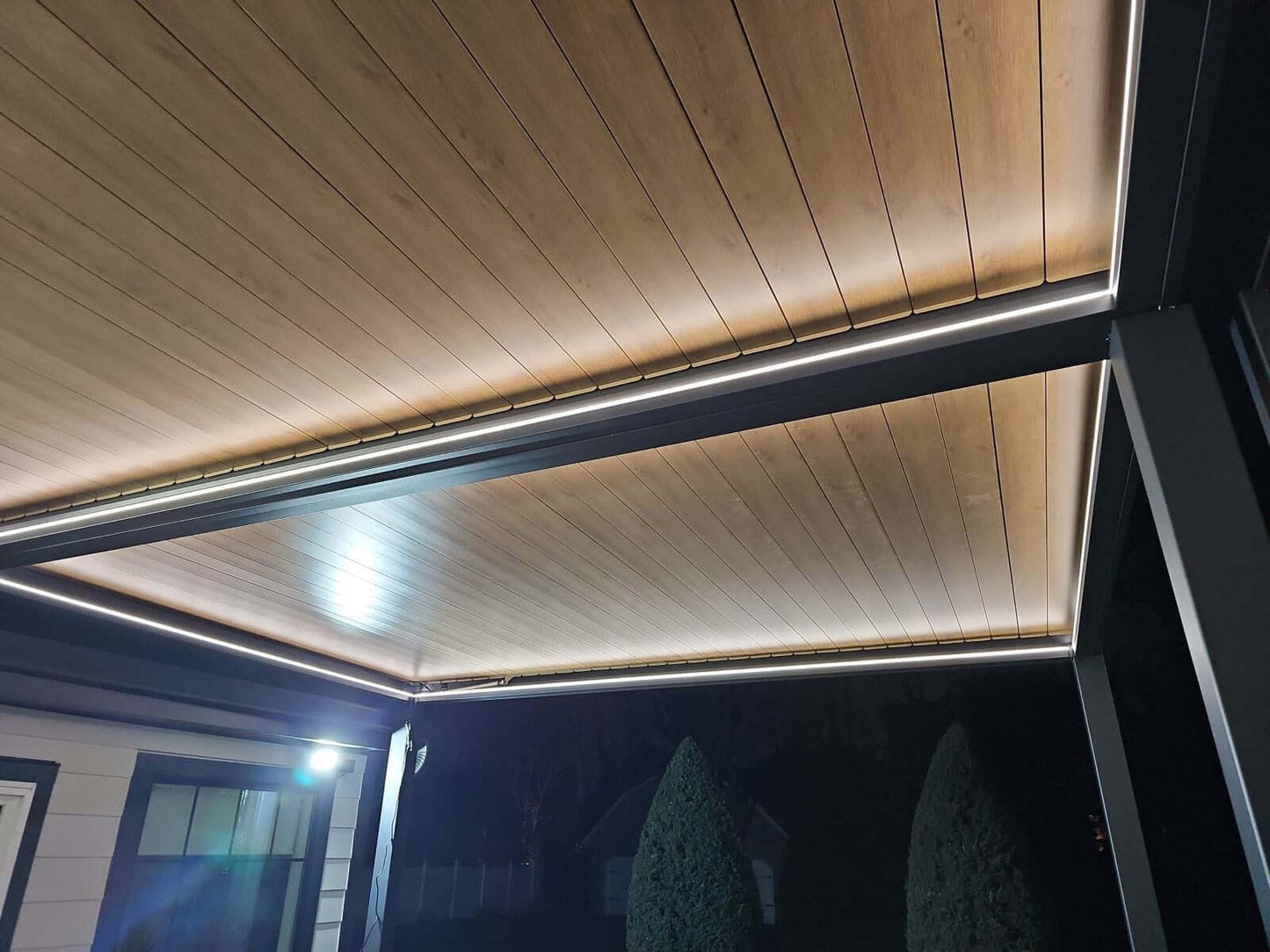 This is a close-up of the pergola roof. The roof is made up of several parallel slats with a wood-like finish, supported by a simple black frame. A white LED strip runs along the inner edge of the roof, casting a bright, even light that illuminates the space below and clearly highlights the pergola’s shape, creating a striking contrast with the dark frame.