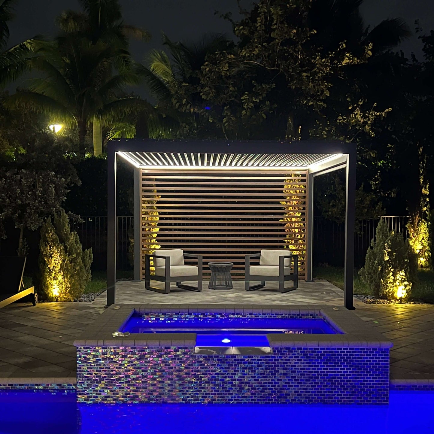 The photo shows a poolside lounge area illuminated by night lighting. The black modern pergola features a motorized louvered roof, with the louvers in an open position. It is equipped with horizontally arranged wood-grain louver panels, adding a decorative touch and creating a semi-enclosed feel. The structure is sleek, made of black columns and beams. Under the pergola, there are two gray modern armchairs and a small gray woven round table positioned between them.