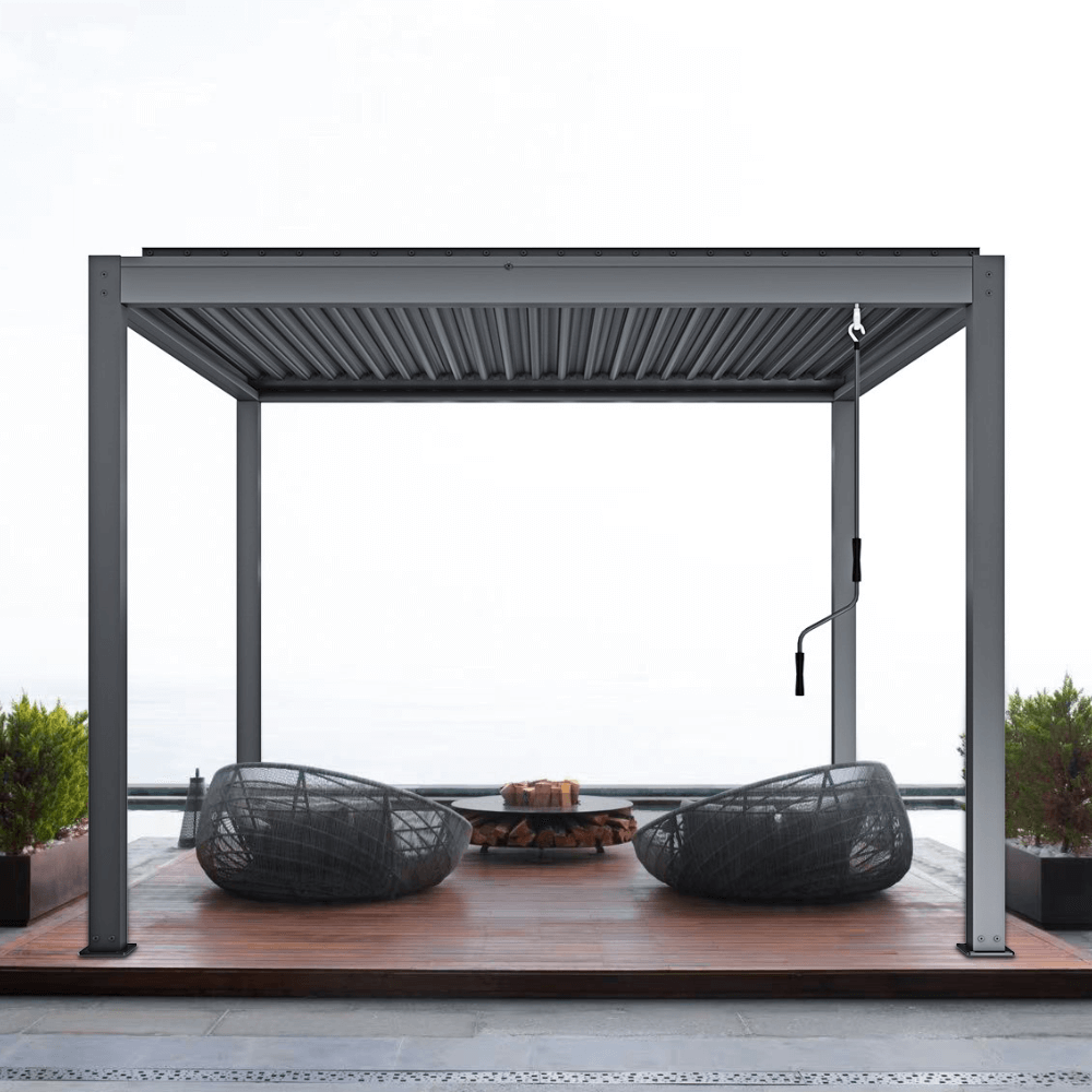 The deep gray Starry Pergola features a manually retractable sunshade, with the canopy partially rolled back, exposing sections of the gray louvered structure above. The pergola's frame is made of sturdy dark gray posts and beams. On the terrace below, two gray woven lounge chairs are placed, along with a round fire pit, its center stacked with firewood, creating a warm and inviting outdoor space.
