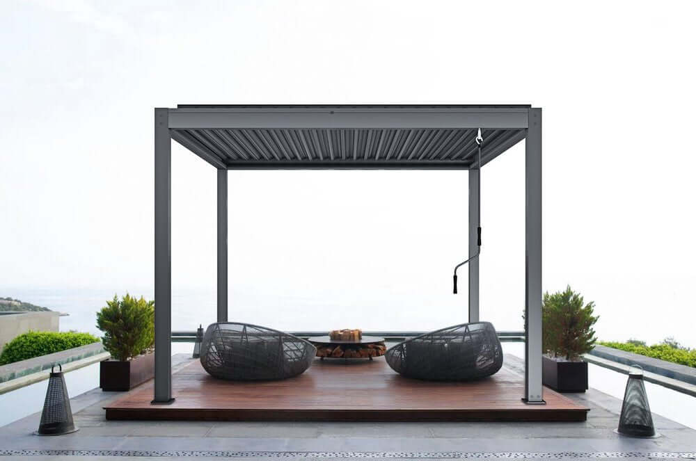 The dark gray Starry Pergola, featuring a manually retractable canopy, stands on a wood-decked terrace with part of its gray louvered structure exposed. Its frame is built from thick dark gray beams and posts in a clean, modern style. On the right side, a hand crank is visible for adjusting the canopy. The terrace is furnished with two gray woven lounge chairs and a round fire pit filled with stacked firewood, adding warmth and charm to the outdoor space.