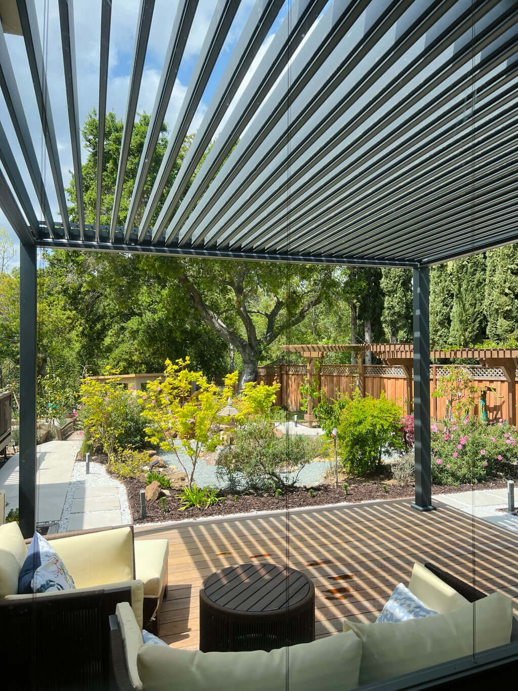 A louvered pergola with open slats frames a lush backyard garden with cream lounge chairs and a wood deck below.