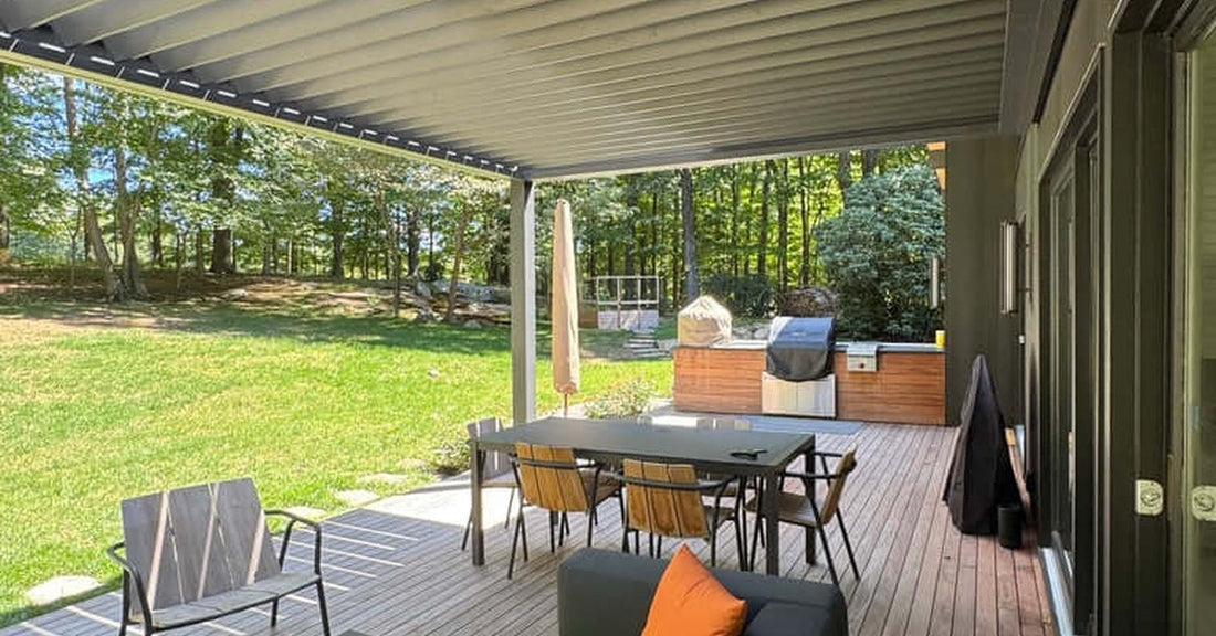 A covered backyard area with a shaded pergola, an outdoor sofa, a dining table, and a grill overlooking a sunny wooded lawn.