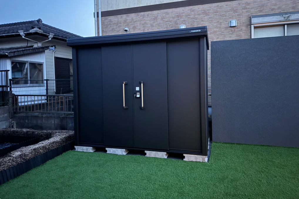 A black metal storage shed with double doors installed on concrete blocks over artificial turf in a residential yard.