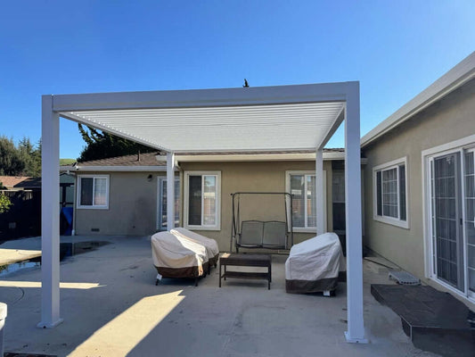 A white aluminum pergola placed in a home’s backyard over three covered chairs next to large sliding doors.