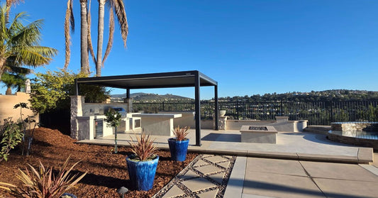 A large patio with a concrete floor and dry mulch with plants, a pergola over an outdoor kitchen next to a fireplace.