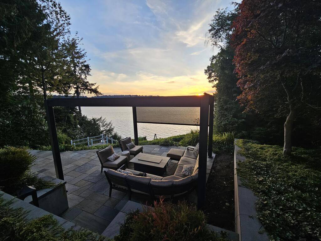 An outdoor living space surrounded by greenery and trees with rock floors, a sectional couch, and a pergola near a lake.