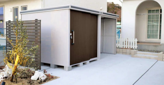 A modern outdoor storage shed with sliding doors, positioned beside a residential home on a concrete patio.