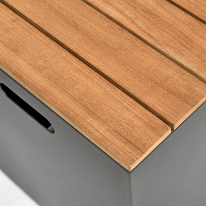 This close-up photo highlights the top and partial side details of the Woodbury Collection Fire Pit storage box. The top is made up of parallel light brown faux wood slats, with small gaps between them that showcase the wood-like texture. The deep gray side panels of the storage box are visible, with the handles designed with smooth, rounded edges.