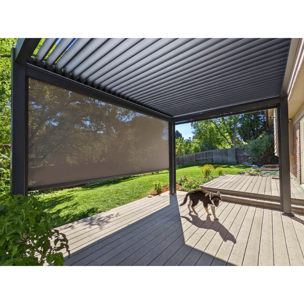 A 13 ft x 19 ft dark gray Bon Pergola with electric louvers, featuring edge lighting, is installed on a wooden deck in the backyard. The louvers are partially open, casting long, narrow shadows. The pergola's structure is made of sturdy dark gray columns and beams. One side of the pergola is equipped with dark sunshades, partially lowered to provide shade and privacy, creating a comfortable outdoor living space.