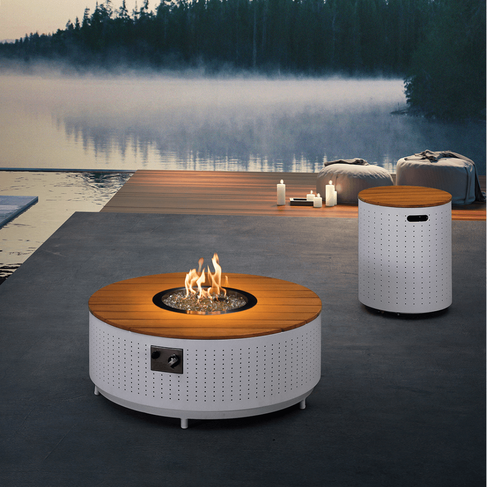 The Cypress Collection Fire Pit features a round fire pit with a white perforated metal base and a light wood slat tabletop. The center holds a circular gas fire pit surrounded by transparent decorative stones, with a metal control panel on the side. Behind it is a cylindrical storage box, matching the table’s base, with a top made of light wood slats and a rectangular handle opening. The setting includes a serene lakeside view at dusk, with a slight mist, creating a peaceful, relaxing ambiance.