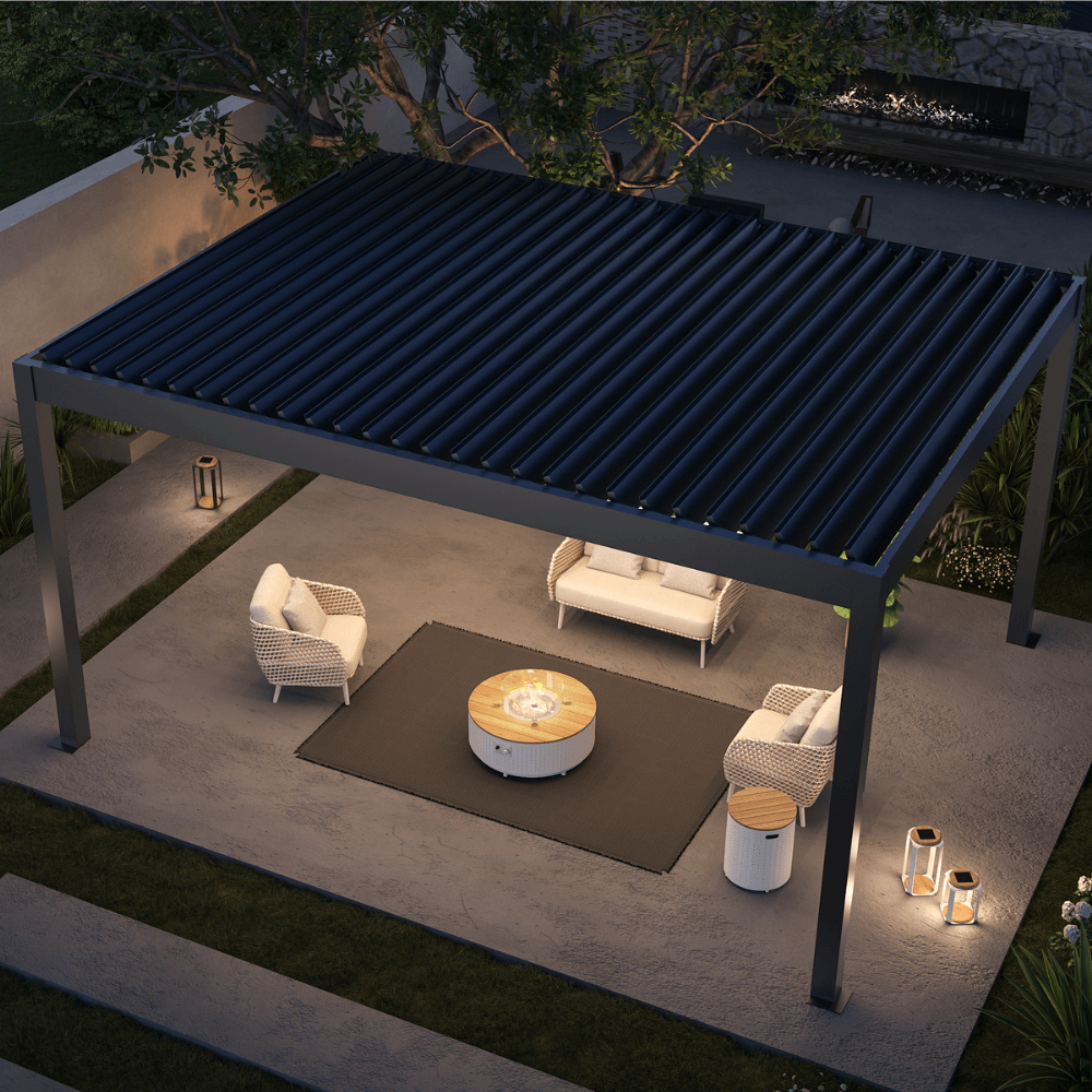 A dark pergola with partially opened louvers features a sturdy frame made of thick dark posts and beams. Beneath, a set of off-white outdoor lounge furniture is arranged, with a white round coffee table at the center. The table holds a gas fire pit, surrounded by a clear glass wind guard. Next to one of the armchairs, there’s a white cylindrical side table, adding a sleek touch to the seating area.