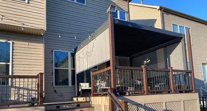 The main structure of the pergola is black, with a retractable top partially rolled back, revealing the louvered structure beneath. One side of the pergola is attached to the exterior wall of the house, while the other is supported by sturdy posts. The side features light-colored motorized blinds, with part of the blinds lowered to provide shade and privacy. The blinds appear to be made of thick material, effectively blocking sunlight.