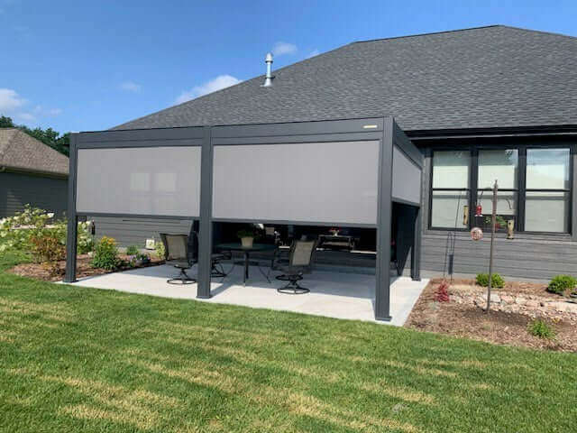 The Villa Pergola features a closed louvered top, creating a sleek black roof. The structure is supported by sturdy black posts and beams. On the sides, light gray motorized blinds are installed, with part of them lowered to provide shade and privacy. The blinds appear to have some translucency, effectively blocking direct sunlight while still allowing light to filter through.