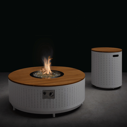This product shot features the Cypress Collection Fire Pit and propane tank storage box. The round coffee table has a white perforated metal base and a light-colored faux wood slat tabletop. In the center is a round gas fire pit with burning flames, surrounded by transparent decorative stones. On the right is a cylindrical storage box made from the same material as the coffee table base, with a light-colored wood slat top and rectangular handle openings on the side.