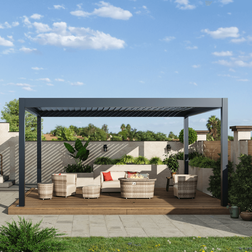 A black motorized louvered pergola with partially opened slats sits atop sturdy black posts and beams. Below, a wood deck hosts a set of beige woven outdoor furniture. At the center, there's a round gas fire pit coffee table, enclosed in a clear glass wind guard. Lush green plants line the wall in front, with a backdrop of tall trees and blue skies, bringing a fresh, natural vibe to the space.