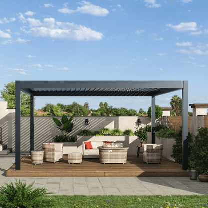 A black motorized louvered pergola with partially opened slats sits atop sturdy black posts and beams. Below, a wood deck hosts a set of beige woven outdoor furniture. At the center, there's a round gas fire pit coffee table, enclosed in a clear glass wind guard. Lush green plants line the wall in front, with a backdrop of tall trees and blue skies, bringing a fresh, natural vibe to the space.