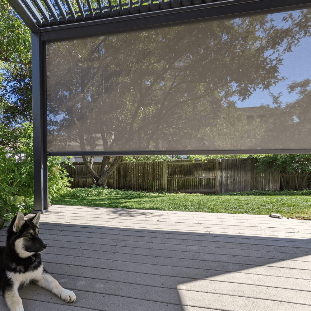 The dark gray Bon Pergola features a partially open louvered roof, casting long, slender shadows across the space below. On one side, a light brown sunshade screen is partially lowered, offering both shade and a bit of privacy. Beneath the pergola lies a patio decked in light gray wood, adding a clean and modern touch to the outdoor setting.