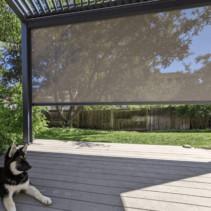 The dark gray Bon Pergola features a partially open louvered roof, casting long, slender shadows across the space below. On one side, a light brown sunshade screen is partially lowered, offering both shade and a bit of privacy. Beneath the pergola lies a patio decked in light gray wood, adding a clean and modern touch to the outdoor setting.